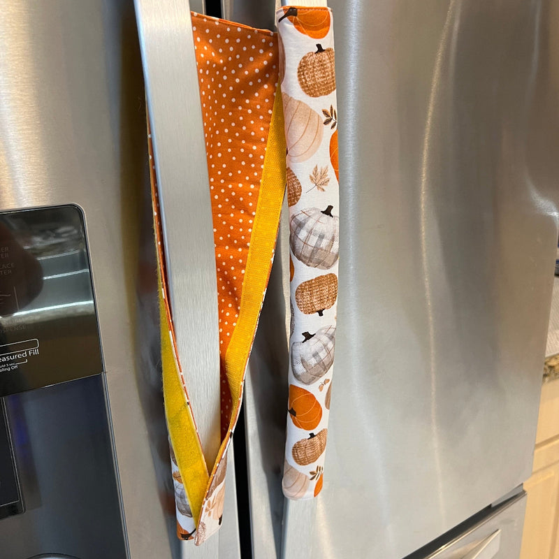 Autumn refrigerator door handle covers hanging on a stainless steel refrigerator. One handle is orange with white polka dots and a yellow border, while the other has a white background with orange, beige, and brown pumpkins. Showing the flip side.