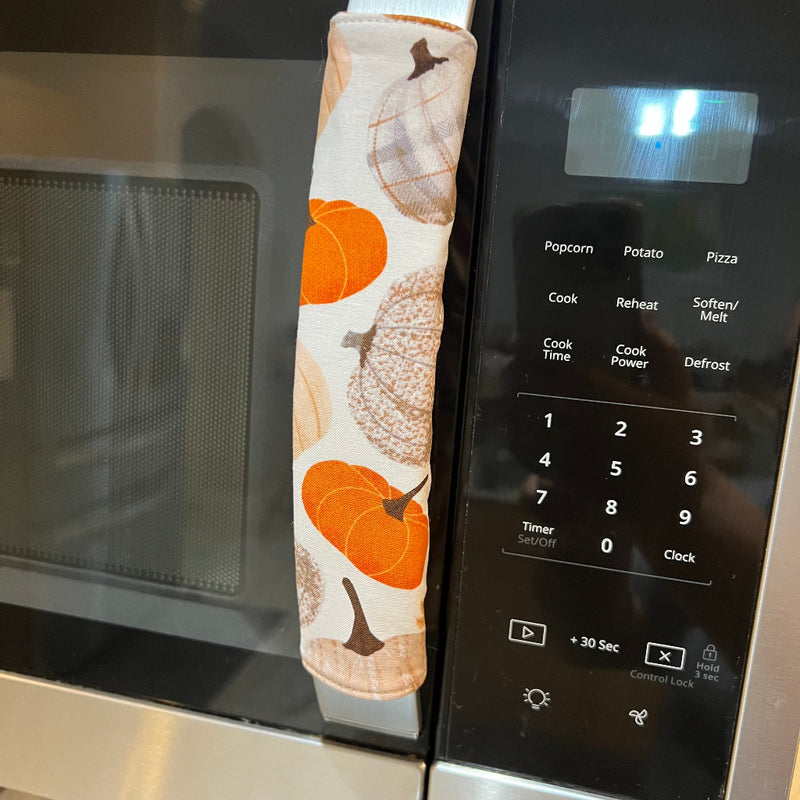 A close-up of a microwave handle covered in a fabric sleeve featuring an autumn pumpkin pattern in orange, beige, and brown on a white background.