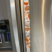 A stainless steel refrigerator handle covered with a vertical fabric sleeve featuring a fall harvest pattern of orange pumpkins, beige pumpkins, and plaid motifs.