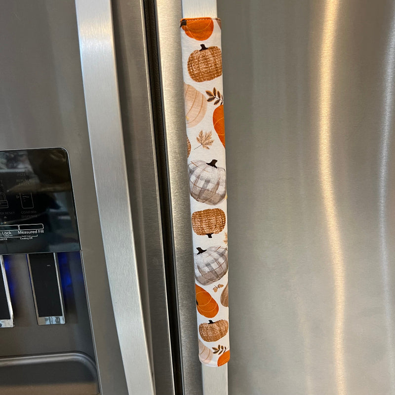 A stainless steel refrigerator handle covered with a vertical fabric sleeve featuring a fall harvest pattern of orange pumpkins, beige pumpkins, and plaid motifs.