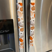 Two white refrigerator handles covered in fabric sleeves with an autumn pumpkin print in orange, beige, and brown, shown on a stainless steel refrigerator.
