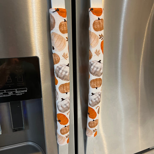 Two refrigerator handles covered in fabric sleeves with an autumn pumpkin print in orange, beige, and brown, shown on a stainless steel refrigerator.