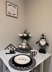 Decorative items on a white surface, including a small house, flowers, and a 'Merry' plate in black and white buffalo plaid.