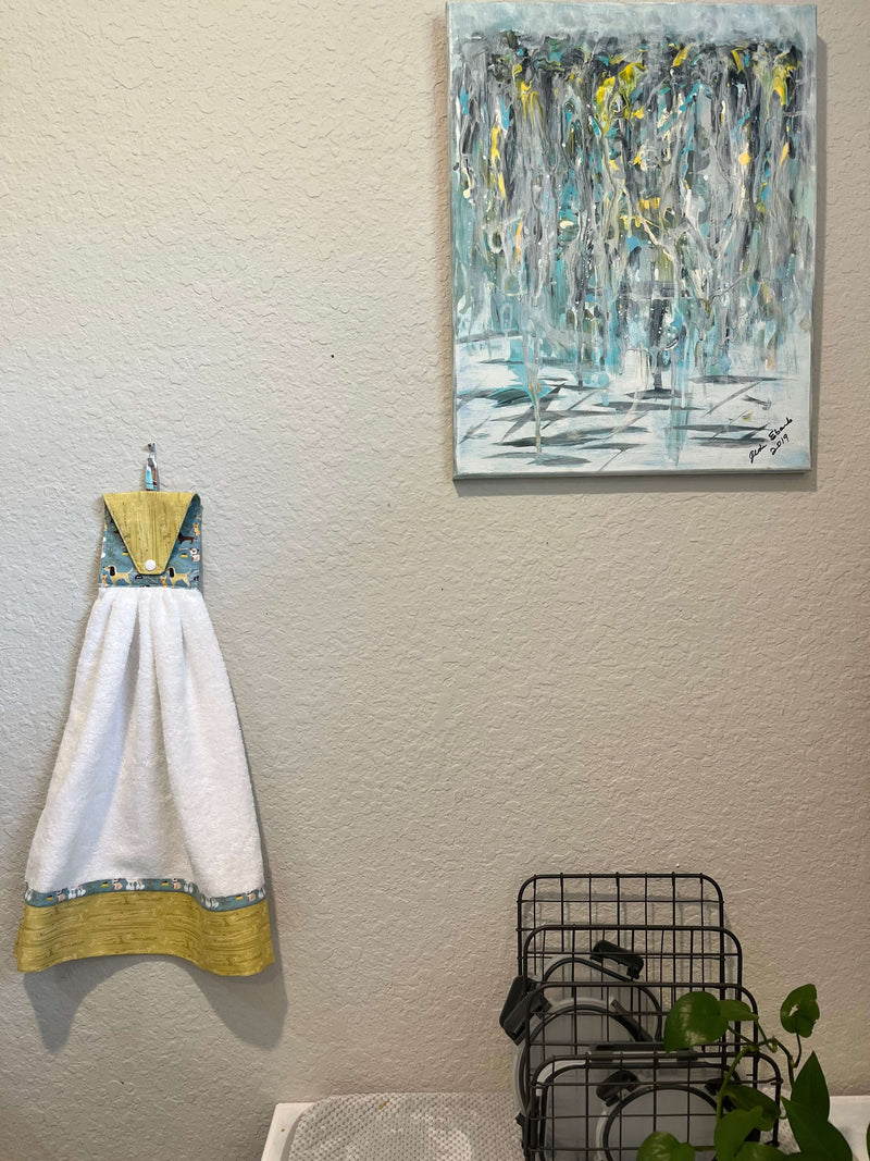 Handmade kitchen towel with a blue and yellow design hanging on a wall beside an abstract painting and a plant.
