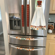 A stainless steel refrigerator with decorative fabric handle covers in a fall harvest pattern, including a white kitchen towel hanging on the right handle.