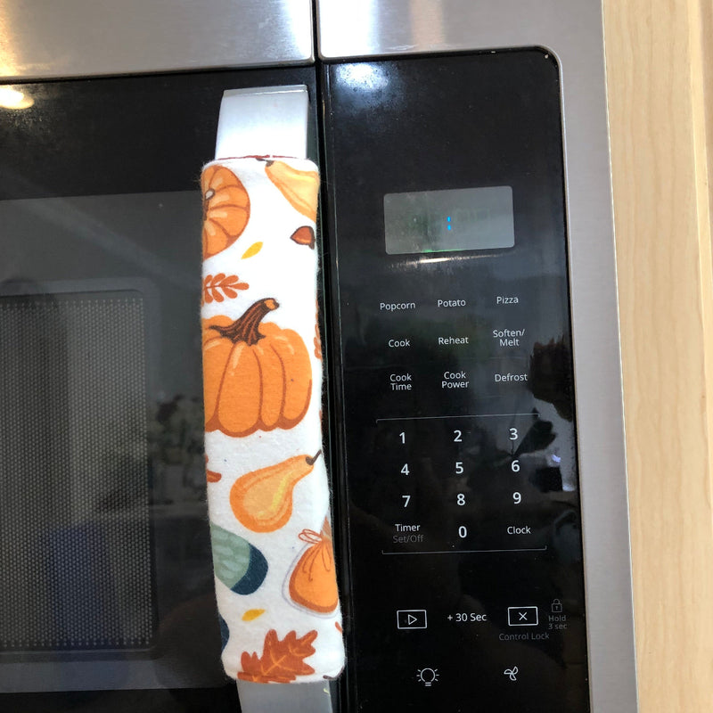 A close-up of a stainless steel microwave with a fabric handle cover featuring an autumn pumpkin and leaf pattern.