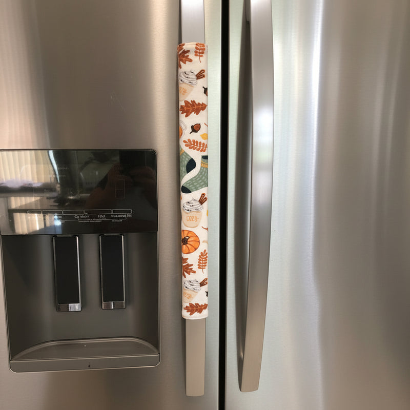 One fabric refrigerator handle coves on a stainless steel fridge. The left handle cover has an autumn-themed pattern with leaves, pumpkins, and a goose on a white background.