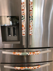 Four stainless steel refrigerator handles covered in matching fabric pull covers with an autumn leaf and pumpkin pattern, installed on a side-by-side refrigerator.