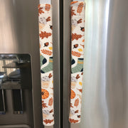 Two stainless steel refrigerator handles covered with vertical fabric pull covers featuring an autumn leaf and pumpkin pattern on a cream background.