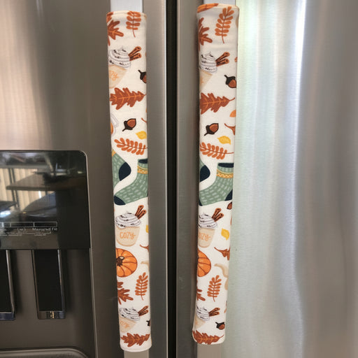 Two stainless steel refrigerator handles covered with vertical fabric pull covers featuring an autumn leaf and pumpkin pattern on a cream background.