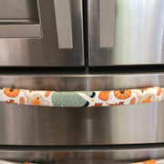 A close-up of a stainless steel refrigerator with two horizontal handles covered in fabric pull covers featuring an autumn pattern of pumpkins, leaves, and a green sweater on an off-white background.