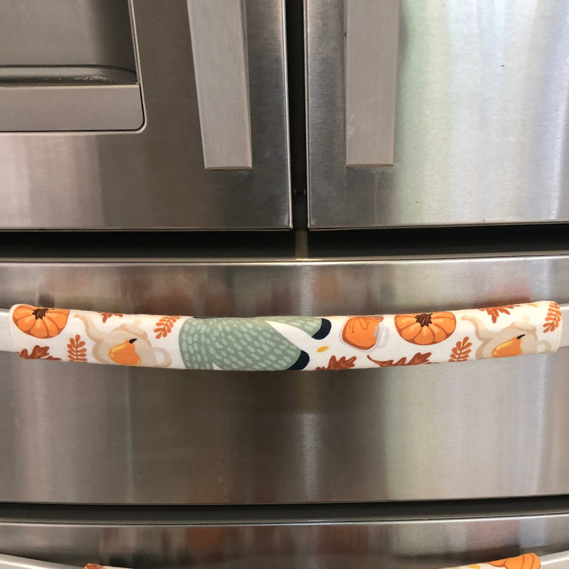 A close-up of a stainless steel refrigerator with two horizontal handles covered in fabric pull covers featuring an autumn pattern of pumpkins, leaves, and a green sweater on an off-white background.