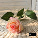 Latex real-touch artificial rose in salmon pink with green leaves on a woven surface.