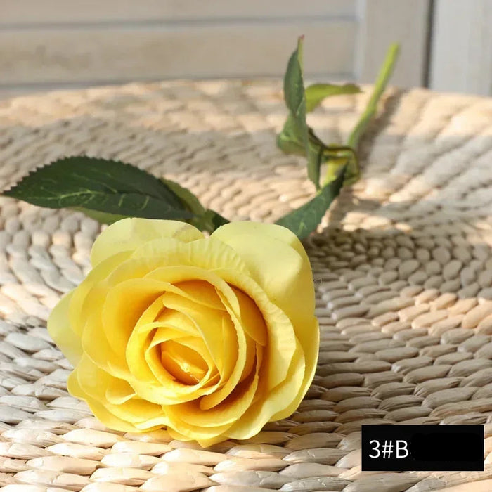 Latex real-touch artificial rose in vibrant yellow color on woven surface with green leaves.