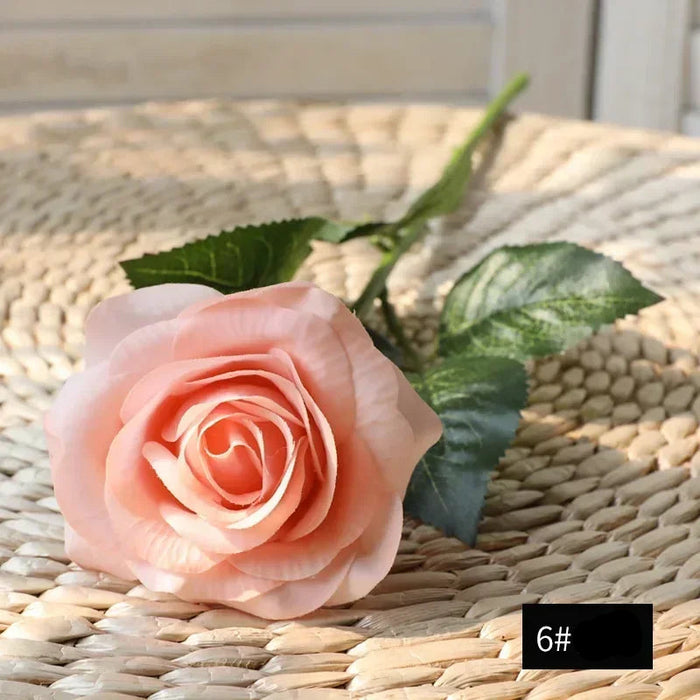 Latex real-touch artificial rose in soft peach color resting on woven surface, highlighting its realistic petals and greenery.