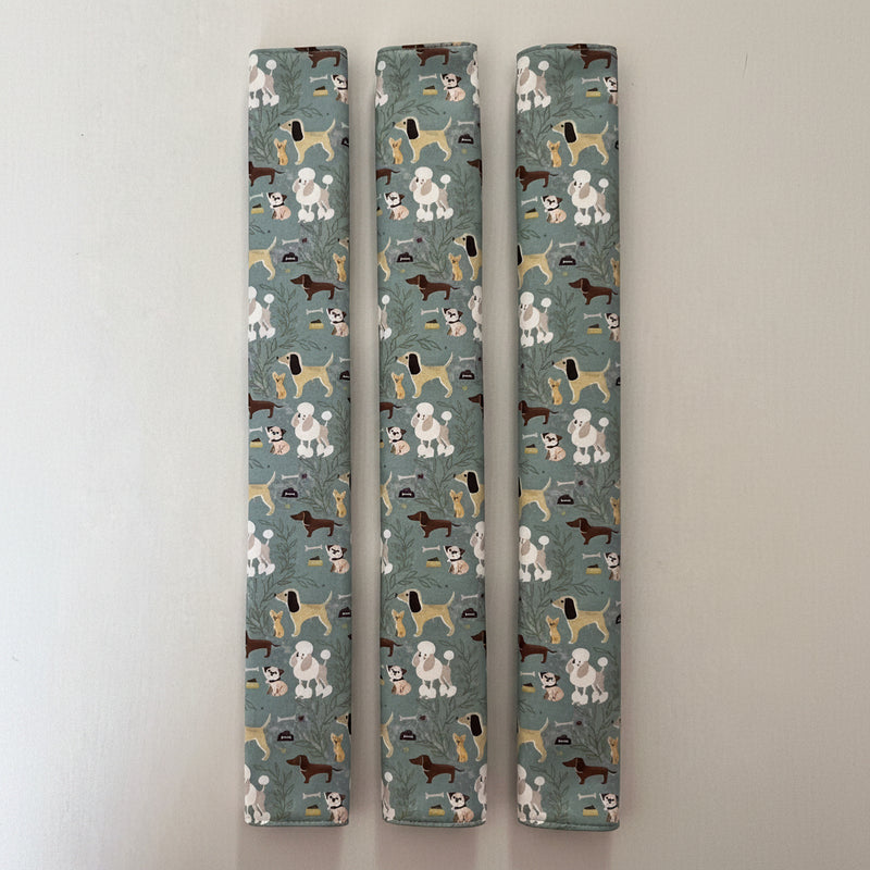 Three fridge handle covers laid vertically side by side on a plain off-white surface, printed with a repeating pattern of various dog breeds in white, brown, tan, and black on a muted blue-green background.