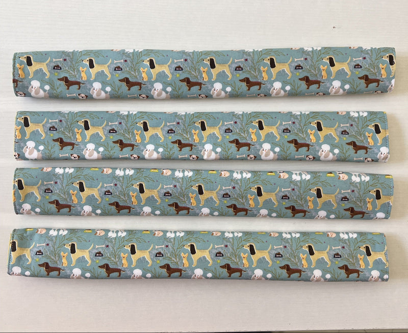 Four fridge handle covers with a repeating pattern of various dog breeds on a muted sage green background, displayed horizontally on a light wood surface.