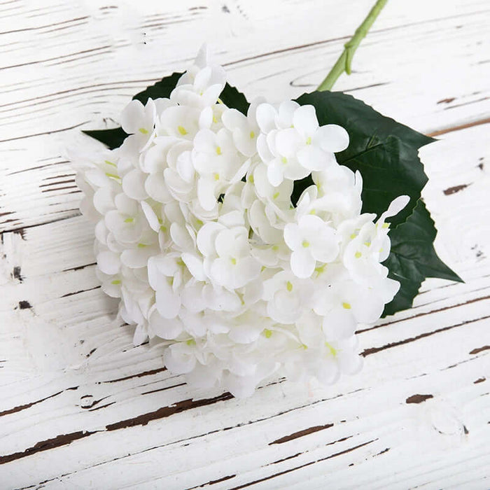 Realistic white Latex Hydrangea artificial flowers with green leaves on a rustic wooden surface.