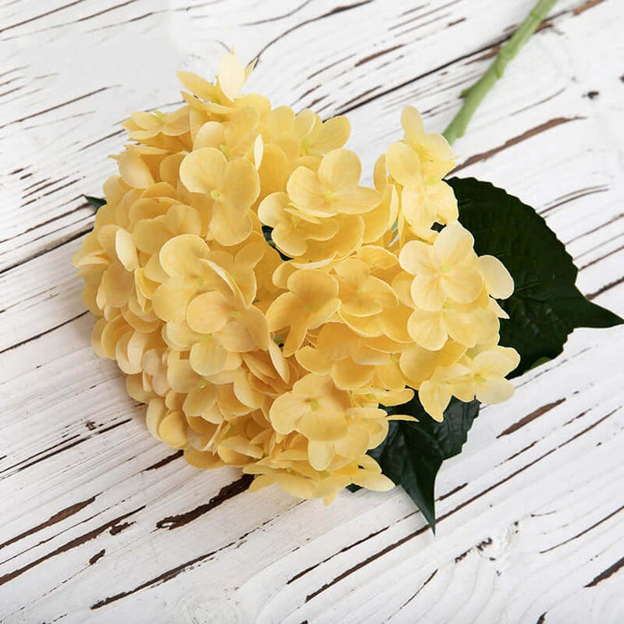 Yellow latex hydrangea artificial flowers with green leaves on a wooden background, realistic and hypo-allergenic.