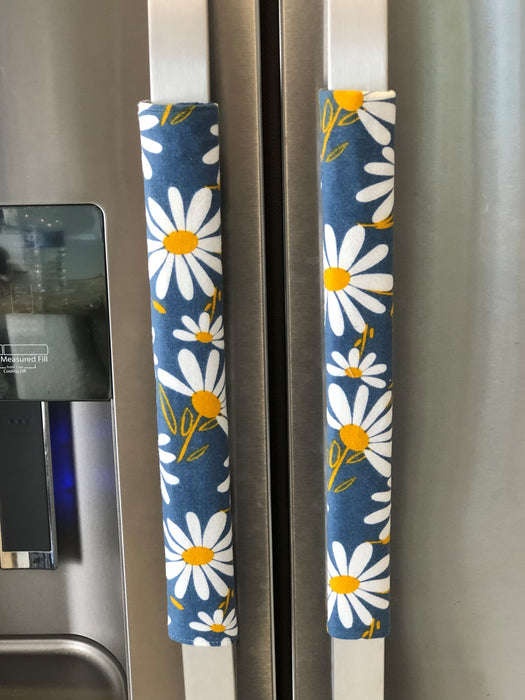 Decorative white and yellow daisy handle covers on blue appliance handles, enhancing kitchen style and protection.