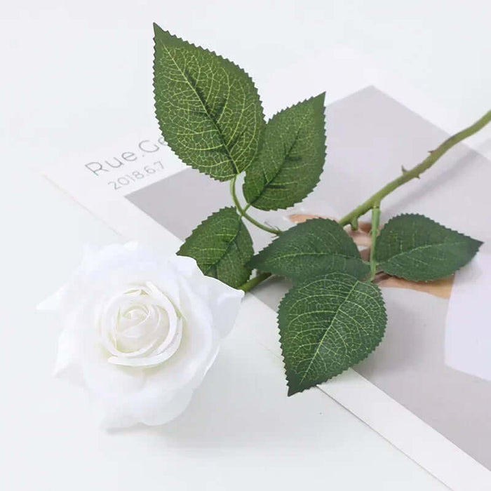Latex real-touch artificial rose with white petals and green leaves on a magazine background.