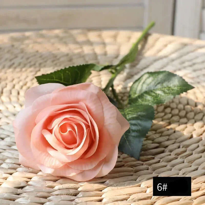 Latex real-touch artificial rose in soft peach color resting on woven surface, highlighting its realistic petals and greenery.